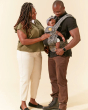 Man and woman stood on a beige background looking down at a baby in a Tula explore baby wearing carrier