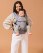Woman stood in front of a brown background carrying a baby in a Tula explore coast beyond baby wearing carrier