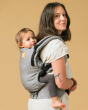 Woman stood on a yellow background carrying a baby in the Tula ash linen baby carrier
