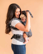 Woman stood on an orange background wearing the Tula eco-friendly free to grow baby carrier in the patchwork checkers print