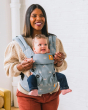 Woman stood in front of a white wall carrying a baby in the Tula blue sky front facing baby carrier