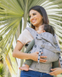 Woman stood under a palm tree with a baby in the Tula adjustable explore baby carrier