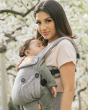 Woman stood under some blossom trees wearing the Tula grey herringbone baby carrier