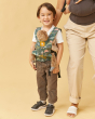 Boy stood on a beige background wearing a Tula mini land before tula toy carrier, next to a woman wearing a grey baby carrier