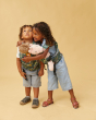 Girl and boy hugging on a beige background, wearing matching Tula mini toy baby carriers with cuddly toys inside