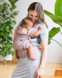 Woman stood in front of some large plants, holding a baby in a Tula explore linen baby carrier