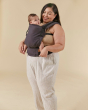 Woman stood on a beige background with a baby in a grey Tula free to grow baby carrier