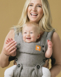 Woman stood in front of a yellow background carrying a toddler in the Tula eco-friendly explore ash baby carrier