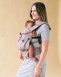 Woman stood on a beige background wearing a Tula linen explore baby carrier in the aubergine colour