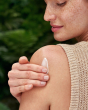 A close up shot of an adult smiling and using the Weleda Skin Foot set cream on the skin of their shoulder