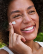 A close up shot of an adult smiling and using the Weleda Skin Foot set cream on their face