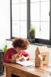Child sat at a wooden table drawing next to a small and large babai eco-friendly wooden dollhouse toy