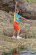 A happy child climbing rocks at the beach, wearing the Frugi Children's Organic Cotton Hotchpotch Applique T-Shirt - Lobster, and orange shorts