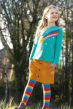Child looking away from camera, wearing the Frugi gold cord Carly button down mini skirt, turquoise teal top with Rainbow and star applique, striped rainbow tights, and boots with trees in the background