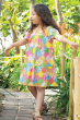 A child walking among tress and grassed in the sun, wearing the Frugi Children's Organic Cotton Shaya Tiered Dress - Patchwork. A beautiful summer dress with three colourful patchwork tiers, a square neck and short puffed sleeves. This colourful children'