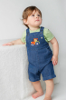 A child looking up and stood in front of a fluffy covered table, wearing the Frugi Organic Carnkie Chambray Denim Dungarees - Tractor and a light green t-shirt undernearth