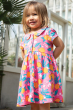 A child smiling in the sun and stood in front of a greenhouse, wearing the Frugi Organic Lettie Dress - Orange Blossom