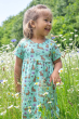 A child happily playing in a flower field full of white and purple flowers, wearing the Frugi Organic Cotton Tallie Dress - Riverine Rabbits. Made with GOTS Organci Cotton, this dress is a beautoful mint green colour, with decorative brown rabbits, yellow