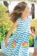 A child happily playing in the sun, wearing the Frugi Children's Organic Cotton Samantha Sleeveless Summer Dress - Wave Stripe / Shell. A gorgeous light blue and white wavy print with two shell applique patches, on a cream background