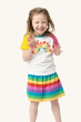 A child happily pulling a funny face whilst wearing the Frugi Children's Organic Cotton Nyomi Raglan T-Shirt - Rainbow. A fun short-sleeved t-shirt for kids from Frugi has a white body, contrasting pink and yellow raglan sleeves, and features a colourful 