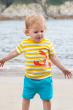 A child happily playing on a beach with the sea/ocean in the background, wearing the Frugi Organic Easy On Outfit - Dandelion Stripe / Lobster