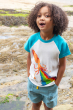 A child happily stood with their hands in their pockets with a seaside background, wearing the Frugi Children's Organic Cotton Robbie Raglan T-Shirt - Squid. A soft 100% organic cotton t-shirt with a white body, contrasting blue raglan sleeves, and featur