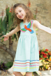 A child smiling, stood with their arms open wide in front of a large leafy green and orange flowering plant, wearing the Frugi Organic Phebe Party Dress - Spring Mint / Macaw.