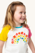 A child smiling happily whilst wearing the Frugi Children's Organic Cotton Nyomi Raglan T-Shirt - Rainbow. A fun short-sleeved t-shirt for kids from Frugi has a white body, contrasting pink and yellow raglan sleeves, and features a colourful floral rainbo