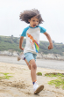A child running along the sand, wearing the Frugi Children's Organic Cotton Robbie Raglan T-Shirt - Squid. A soft 100% organic cotton t-shirt with a white body, contrasting blue raglan sleeves, and features a playful squid shooting out rainbow ink appliqu