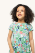 A child looking off into the distance happily, and wearing the Frugi Children's Organic Cotton Spring Skater Dress - Tropical Birds. A beautiful tropical bird and flower print on a twirly short sleeve skater dress, on a cream background
