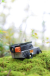 Candylab Drifter Kodiak, a 4x4 off-roader wooden car toy, in grey and black, with metal roof rack and rubber tyres, on mossy, rocky ground with out of focus trees in background, right side rear view of toy