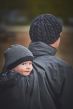 Mamalila Shelter Men's Babywearing Rain Jacket in Anthracite. Rear lifestyle view of this men's technical babywearing rain coat with the baby carrier insert on back. Blurred background