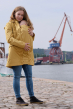 Mamalila Dublin Babywearing Rain Coat in Mustard. A sustainably made babywearing rain coat in mustard yellow. Front lifestyle view, with baby carrier insert at front. Crane and docks in background. 