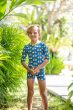 child standing among trees and greenery, smiling wearing the Maxomorra Rainbow Swim Trunks with a matching swim top
