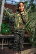 Meyadey Marvellous Macaw, organic, zipped hoodie in rich green with macaw and leaves repeat print. Worn by a young person with dark green trousers surrounded by plants