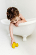 Baby in a small bath tub holding reaching for a yellow Oli & Carol Elvis The Duck Bath Toy which is on the floor