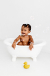 Baby in a small bath tub holding with bubbles floating around with a yellow Oli & Carol Elvis The Duck Bath Toy on the floor in front