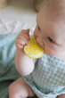 Oli & Carol John Lemon natural rubber teether, being held and chewed on by a pale baby in blue gingham
