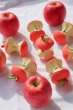 7x Oli & Carol Pepa the Apple natural rubber teether, laid in a circular pattern with fresh red apples around the outside, on white fabric