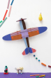 The huge Studio ROOF Deluxe Collection Propellor Plane, a cardboard model plane with geometric patterns and bright colours, put together, on white background wall with geometric shames below and above. Purple surface below.