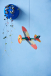 The Studio ROOF Propeller Plane, a cardboard model plane with geometric patterns and bright colours, put together, hanging from ceiling, blue wall behind with a blue wall plant pot containing dangling plant in top left corner.