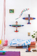 The Studio ROOF Deluxe Condition Glider Plane, a huge cardboard model plane with geometric patterns and bright colours, put together, on a white wall in a bedroom with the other Studio ROOF deluxe plane, above a Childs bed. some geometric shapes in differ