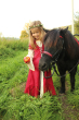 Child wearing the Vah Red Little Marian costume Dress with a cream coloured floral crown feeding an apple to a pony