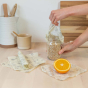 Close up of hands wrapping a glass jar and an orange in the small Abeego plastic-free beeswax food wraps on a kitchen counter