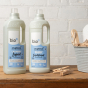 Bio-D fragrance free fabric conditioner and laundry liquid 1 litre bottles on a white brick background and wooden table, with a peg basket and pegs next to the bottles