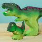 Wooden toy baby T-Rex in vibrant shades of green and purple. The baby T-Rex stands upright in a forward stance. The toy has painted eyes, claws, and body detail. The baby T-Rex stands next to an adult version of the toy for a reference of scale.