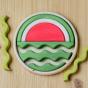 A round wooden puzzle depicting a watermelon. Different shapes fit together to create the final image, the puzzle is partly completed with the remaining shapes shown alongside. The base of the puzzle has the shape template printed on it.