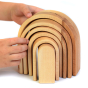 Bumbu Natural Wooden Stacking Arches. The toy sits on a white surface an adult hand is shown to denote scale.