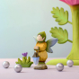 The Bumbu Wooden Winged Elf holding their Wooden lamp and stood next to a small Bumbu grass with a purple flower and small silver balls, with a Large Pink flower in the background 