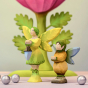 The Bumbu Wooden Winged Elf and Bumbu Woodland Fairy holding the Wooden lamp and stood next to small silver balls, with a Bumbu Large Pink flower in the background 
